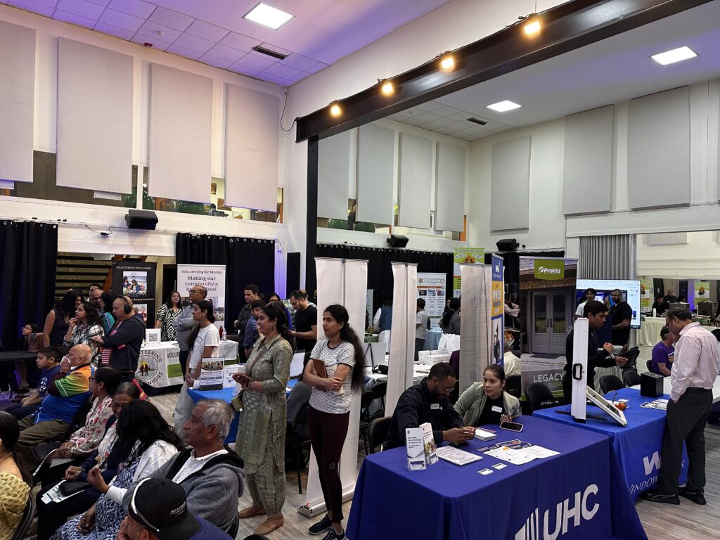 Audience members from seniors to children watch what's happening on stage while in the background community organizations tabling are sharing info and resources to community members