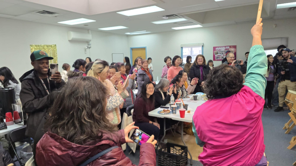 Community members celebrate and gather around a drum performance with cameras in hand and smiles all around